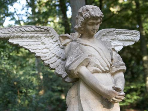 Skulptur eines Engels mit gefalteten Händen und großen Flügeln, umgeben von grünem Wald.